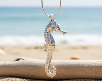 May include: A silver seahorse pendant necklace. The seahorse is detailed with textured scales and fins, suspended from a delicate silver chain. The background shows a sandy beach, driftwood, and the ocean.