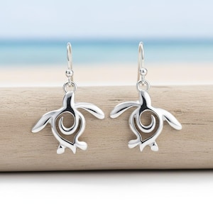 May include: A pair of silver-colored earrings featuring a sea turtle design. Each earring has a turtle shape with a spiral detail in the center and a hook for the ear. The earrings are displayed on a wooden surface with a blurred ocean background.