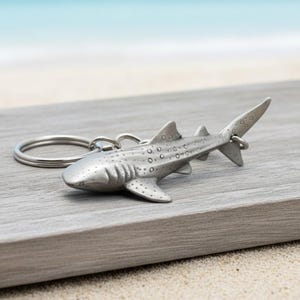 May include: A silver-colored whale shark keychain. The keychain features a detailed, three-dimensional design of a whale shark with visible spots and fins. The shark is attached to a silver key ring, perfect for ocean lovers.