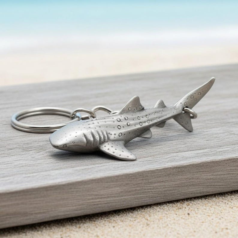 May include: A silver-colored whale shark keychain. The keychain features a detailed, three-dimensional design of a whale shark with visible spots and fins. The shark is attached to a silver key ring, perfect for ocean lovers.
