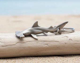 May include: A silver-colored hammerhead shark keychain. The shark is detailed with fins, gills, and a tail, attached to a silver keyring. The keychain rests on a piece of driftwood, with a sandy beach and ocean in the background.