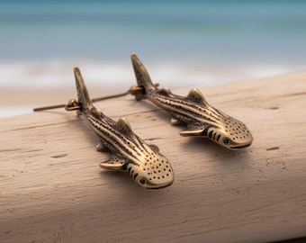 May include: A pair of bronze-colored earrings shaped like whale sharks. The earrings have detailed fins, stripes, and dotted patterns. The earrings are displayed on a piece of driftwood with a blurred ocean background.