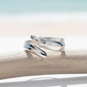 Op de afbeelding: Een zilverkleurige ring in de vorm van een slang, opgerold. De kop van de slang is gedetailleerd met schubben en een kleine edelsteen. De ring wordt gepresenteerd op een stuk drijfhout, met een vage achtergrond van strand en lucht.