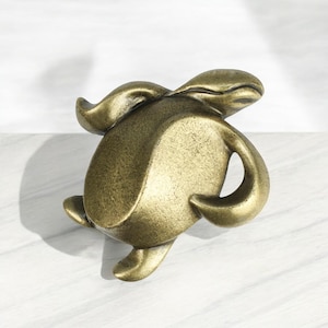 May include: A gold-toned metal turtle figurine with a detailed, textured surface. The turtle is in a walking position with its head turned to the side.