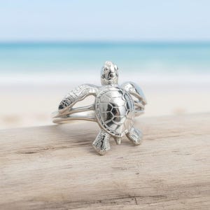May include: A silver-colored ring featuring a detailed sea turtle design. The ring is set against a blurred background of a beach and ocean. The turtle's shell and flippers are intricately crafted, showcasing a nautical theme.