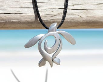 May include: A silver-colored turtle pendant necklace with a black cord. The pendant features a stylized turtle design with a swirling pattern in the center. The background shows a beach scene with a wooden beam.