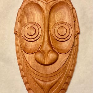 May include: A wooden mask with a carved face, featuring large eyes, a wide mouth, and a detailed design. The mask is a rich brown colour and has a smooth, polished finish.