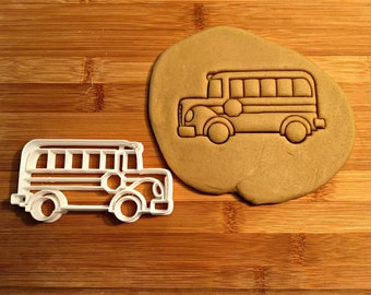 May include: A white plastic cookie cutter in the shape of a school bus. The cutter is on a wooden surface next to a brown cookie dough cutout of a school bus.