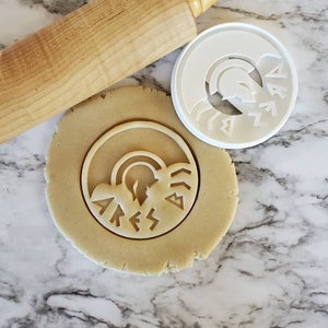 Op de afbeelding: Een witte plastic koekjesvormer met een ontwerp van een Griekse godenhelm en het woord "ARES" in oude Griekse schrift. De koekjesvormer ligt op een marmeren oppervlak naast een deegroller en een cirkel van koekdeeg.