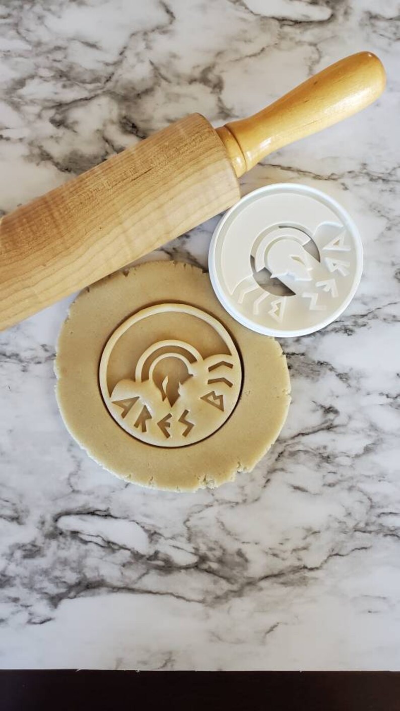 Op de afbeelding: Een witte plastic koekjesvormer met een ontwerp van een Griekse godenhelm en het woord "ARES" in oude Griekse schrift. De koekjesvormer ligt op een marmeren oppervlak naast een deegroller en een cirkel van koekdeeg.
