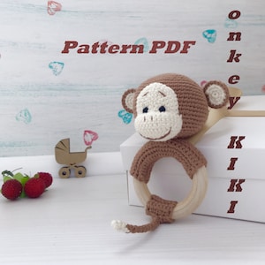 Peut inclure: Hochet en crochet en forme de singe avec un anneau en bois. Le singe est brun avec un visage blanc et une queue brune. Le jouet est sur une boîte blanche avec les mots "Pattern PDF" et "Monkey Kiki" écrits dessus.