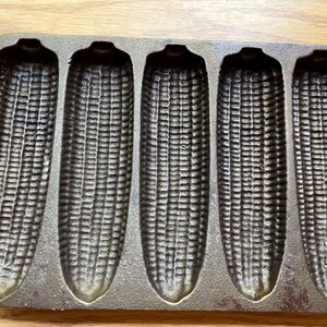 May include: Cast iron corn on the cob baking pan with six individual molds. The pan is dark gray and has a textured surface.