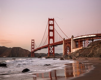 May include: A red suspension bridge with a large archway, spanning a body of water. The bridge is reflected in the water, which is calm and still. The sky is a light blue color, and there are a few birds flying in the air.