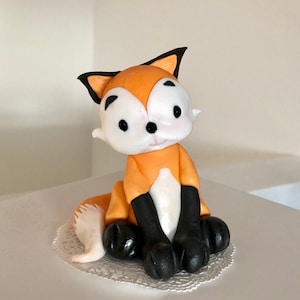 Peut inclure: Une petite figurine de renard orange et blanc avec des oreilles, des yeux et des pattes noirs. Le renard est assis sur un doily en dentelle blanche.