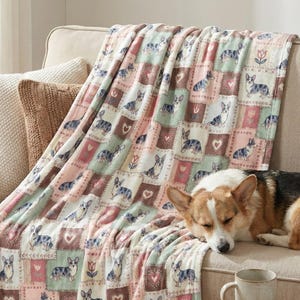 Puede incluir: Una manta suave, de estilo patchwork, sobre un sofá beige. La manta presenta un patrón repetido de perros corgi, corazones y diseños florales en tonos de rosa, verde y marrón. Un perro corgi descansa sobre el sofá.