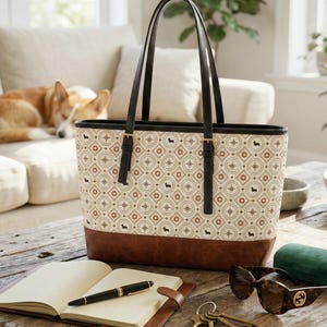 May include: A tote bag with a patterned design featuring brown and black dachshund silhouettes and compass rose motifs. The bag has black handles and a brown leather base. A notebook, pen, sunglasses, and keys rest on a wooden table.