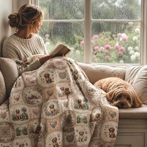 Puede incluir: Una manta beige con un patrón repetitivo de pequeños cachorros marrones, arreglos florales y formas de corazón. La manta está sobre una persona que lee un libro, con un perro de color dorado descansando sobre la tela a su lado.