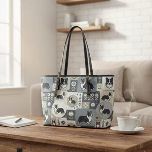 May include: A tote bag featuring a repeating pattern of black and white Corgi dogs in various poses, interspersed with floral and geometric shapes in shades of gray, white, and black. The bag has two black shoulder straps and a rectangular base.