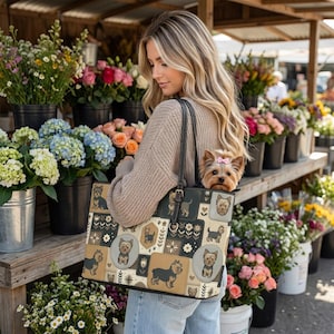 May include: A beige tote bag featuring a repeating pattern of Yorkshire Terrier illustrations, floral motifs, and geometric shapes, with a small Yorkshire Terrier with a pink bow sitting inside the bag, which has black leather shoulder straps.