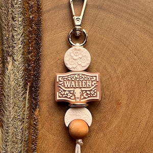 May include: A gold-coloured keychain featuring a white floral-embossed disc, a rectangular charm with the name "WALLEN" and a bull skull design, and a wooden bead. The keychain has a white tassel and a gold clasp.
