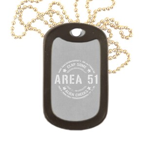 May include: A silver dog tag necklace with a black chain. The dog tag has a black border and a white circle with the text "Clap Some Alien Cheeks Area 51" printed on it.