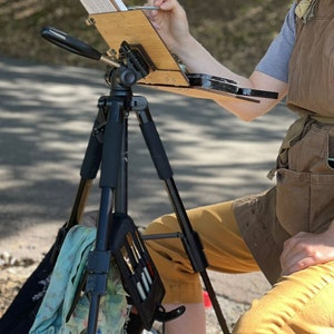 May include: A person painting outdoors with a portable easel set up on a tripod. The easel is made of wood and has a black metal frame. The person is wearing a brown apron and khaki pants. A green cloth is hanging from the tripod.