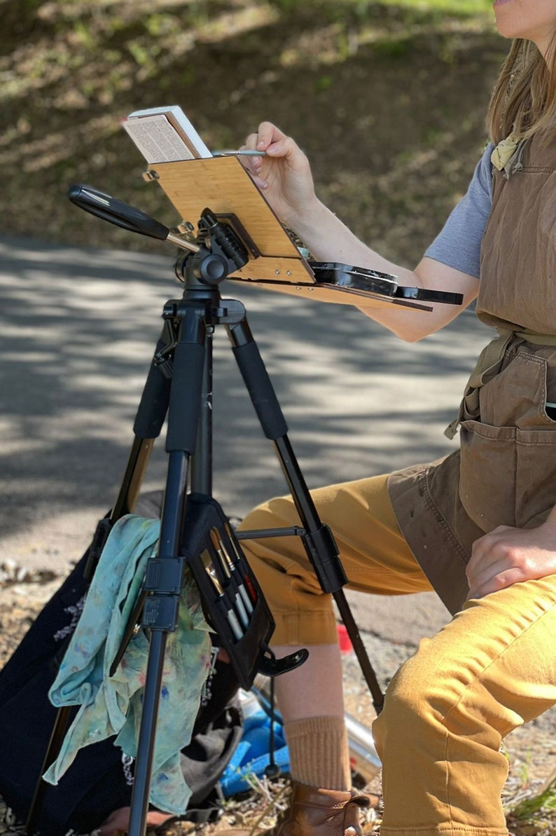 May include: A person painting outdoors with a portable easel set up on a tripod. The easel is made of wood and has a black metal frame. The person is wearing a brown apron and khaki pants. A green cloth is hanging from the tripod.