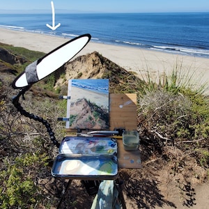 May include: An artist's easel set up on a cliff overlooking the ocean. The easel has a canvas, a paint palette, and a water container. A 12x12 inch  Wimberley Plamp II  compactable light diffuser is positioned above the canvas.