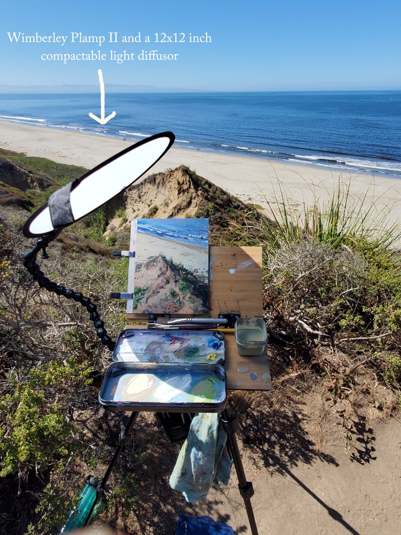 May include: An artist's easel set up on a cliff overlooking the ocean. The easel has a canvas, a paint palette, and a water container. A 12x12 inch  Wimberley Plamp II  compactable light diffuser is positioned above the canvas.