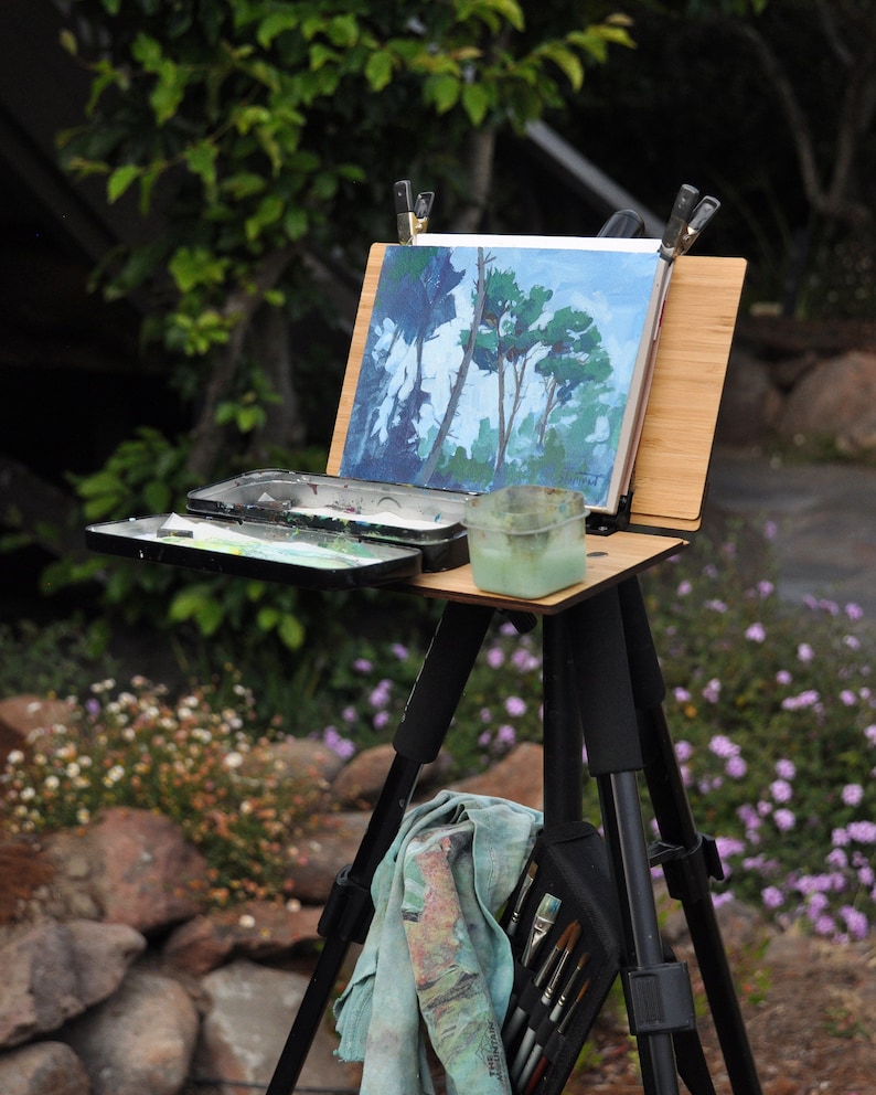 May include: A black easel with a wooden top and a canvas with a painting of trees. The easel is set up outdoors on a grassy surface with a blue cloth hanging from it.