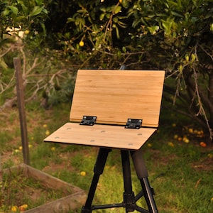 May include: A wooden easel with a bamboo surface, set up on a black tripod in a grassy outdoor setting.