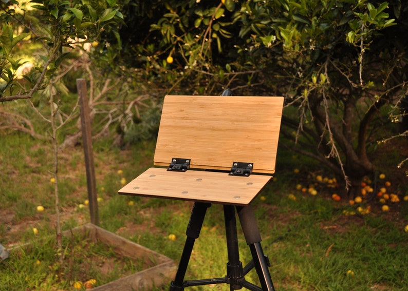 May include: A wooden easel with a bamboo surface, set up on a black tripod in a grassy outdoor setting.