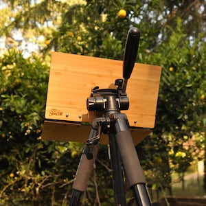 May include: A wooden easel with a black tripod stand. The easel is made of bamboo and has a metal handle. The easel is set up outdoors in front of a green tree.