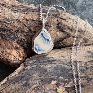 May include: A silver necklace featuring a pendant with a unique, irregular shape. The pendant has a white and blue design, set in a silver bezel. The chain is also silver, and the necklace is displayed on a piece of driftwood.