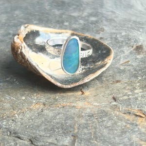 May include: A silver ring with an oval, iridescent blue opal gemstone. The ring is set in a simple band with etched floral designs. The ring is displayed inside a seashell, resting on a gray, textured surface.