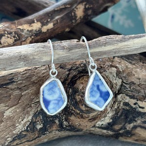 May include: A pair of silver dangle earrings. Each earring features a blue and white patterned, irregular-shaped ceramic piece set in a silver bezel. The earrings hang from silver hooks, showcasing a nautical theme.
