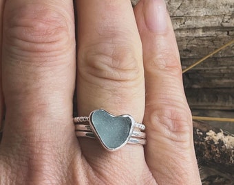 Puede incluir: Un anillo de plata con una pieza de vidrio de mar en forma de corazón engastada en el centro. El anillo está apilado con otras dos bandas de plata.