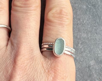 Puede incluir: Un anillo de plata con una piedra de vidrio de mar azul claro engastada en el centro. El anillo está apilado con otras dos bandas de plata.