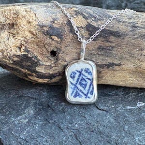 May include: A silver pendant necklace featuring a rectangular, blue and white patterned ceramic piece. The pendant is set in a silver bezel and hangs from a delicate silver chain. The necklace rests on a piece of driftwood.