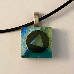 May include: A square glass pendant necklace with a black circle containing a green triangle. The pendant has a blue, green, and iridescent background.