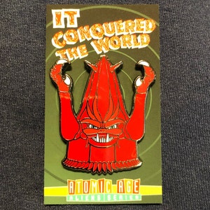 May include: A red and gold enamel pin featuring a monster with the text "IT CONQUERED THE WORLD" and "ATOMIC AGE ALIENS SERIES".