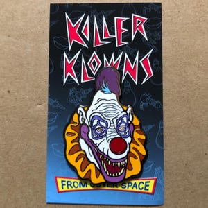 May include: A colourful enamel pin featuring a cartoon clown with a purple and blue mohawk, a white face, red nose, and a toothy grin. The pin is on a card with the text "Killer Klowns from Outer Space".