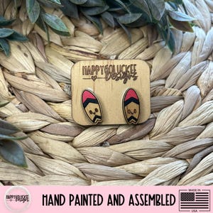May include: Wooden stud earrings featuring a surfboard design with red, black, and tan accents. The earrings have a cute face design. The earrings are hand painted and assembled. The earrings are on a wooden background.