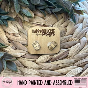 May include: Wooden stud earrings featuring a bandage design. The earrings are on a small wooden square with the text "HAPPYGOLUCKEE Designs". The image also includes the text "HAND PAINTED AND ASSEMBLED" and a small American flag.