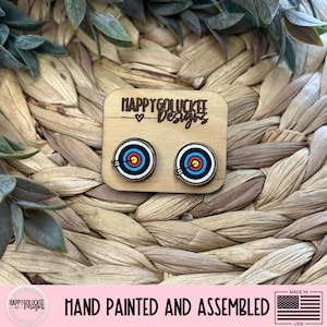 May include: Wooden stud earrings featuring a target design with red, yellow, blue, and white concentric circles. The earrings are mounted on a small wooden card with the text "HAPPYGOLUCKEE Designs". The text "HAND PAINTED AND ASSEMBLED" is also visible.