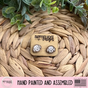 May include: Wooden stud earrings featuring a white and brown dog face design. The earrings are on a small wooden display with the text "HAPPYGOLUCKEE Designs". The image also includes the text "HAND PAINTED AND ASSEMBLED" and a small American flag.