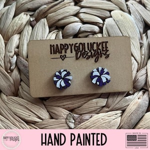 May include: Hand-painted stud earrings with a pom-pom design in white and purple. The earrings are displayed on a brown card with the text "HAPPYGOLUCKEE Designs". The background is a woven texture.