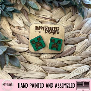May include: Wooden stud earrings featuring a green pixelated design on a square shape. The earrings are displayed on a wooden plaque with the text "HAPPYGOLUCKEE Designs". The bottom of the image reads "HAND PAINTED AND ASSEMBLED".
