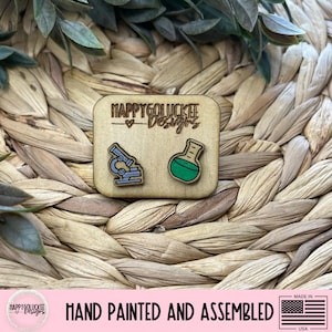 May include: A wooden square featuring a microscope and a flask, hand-painted in blue and green. The text "HAPPYGOLUCKEE Designs" is above the images. The words "HAND PAINTED AND ASSEMBLED" are below the square, with a small American flag.