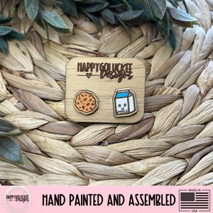 May include: Wooden cookie and milk carton earrings. The earrings are hand-painted and assembled. The earrings are on a wooden square with the text "HAPPYGOLUCKEE Designs". The earrings are made in the USA.